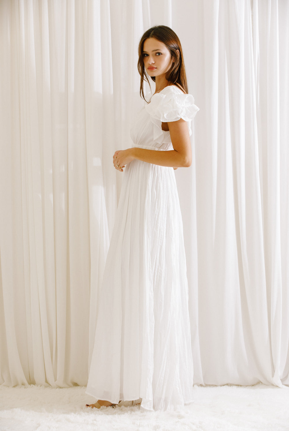 Side view of a woman wearing a white maxi dress standing against a white curtain