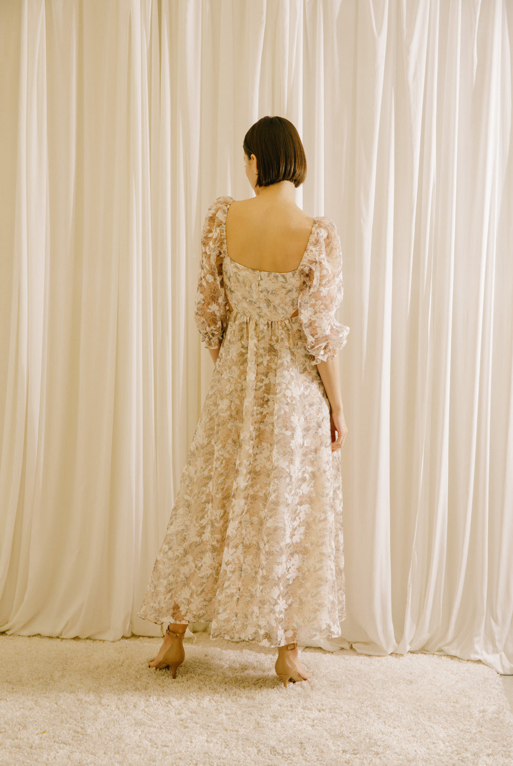 Back view of a model in a floral dress standing against a beige curtain backdrop