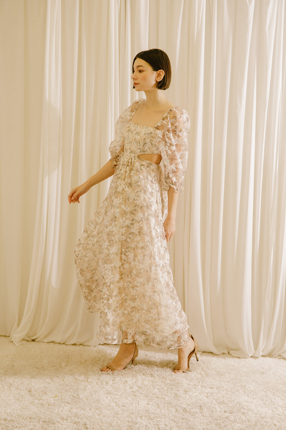 Side view of a model in a floral maxi dress with side cut-outs standing against a beige curtain backdrop