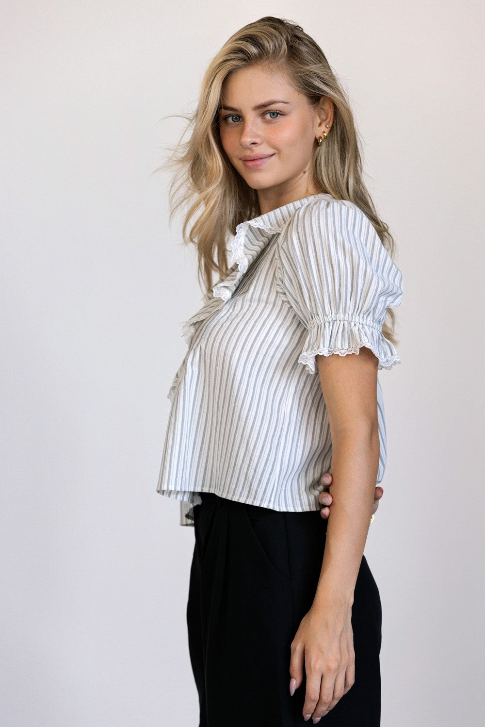 Side view of a woman wearing a striped blouse with ruffled sleeves and black trousers against a plain background