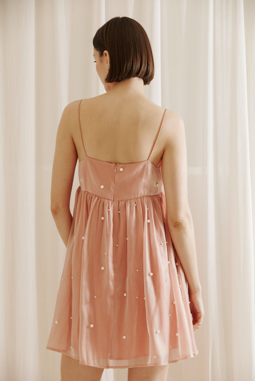 A back view of a person wearing a pink dress with pearl embellishments and an invisible back zipper against a beige curtain background