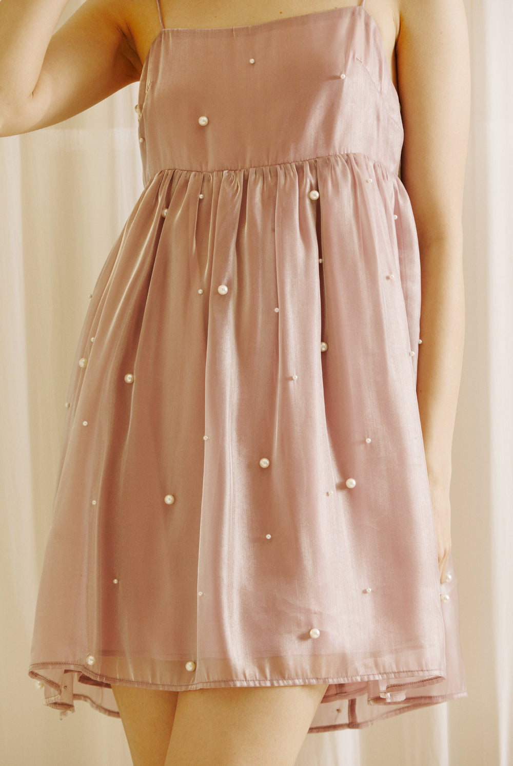 A person wearing a mauve mini dress with pearl embellishments against a beige curtain background