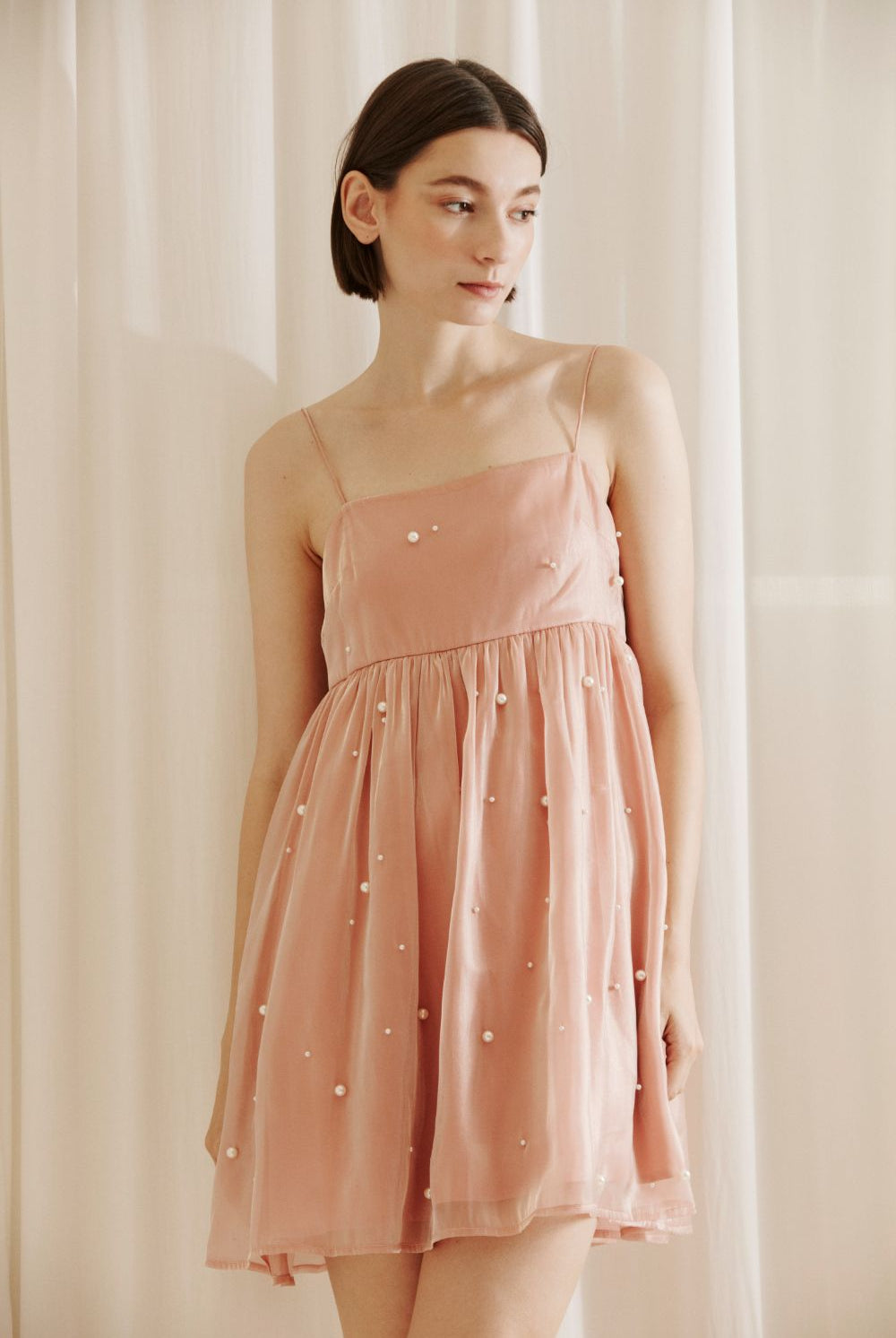 Woman wearing a pink dress with pearl embellishments against a white curtain background