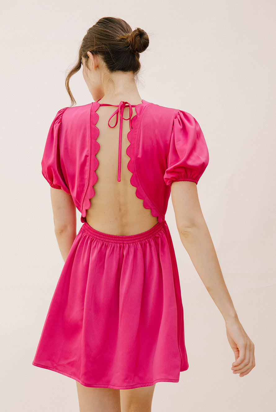 Back view of a woman wearing a bright pink dress with an open back and scalloped hem against a plain background