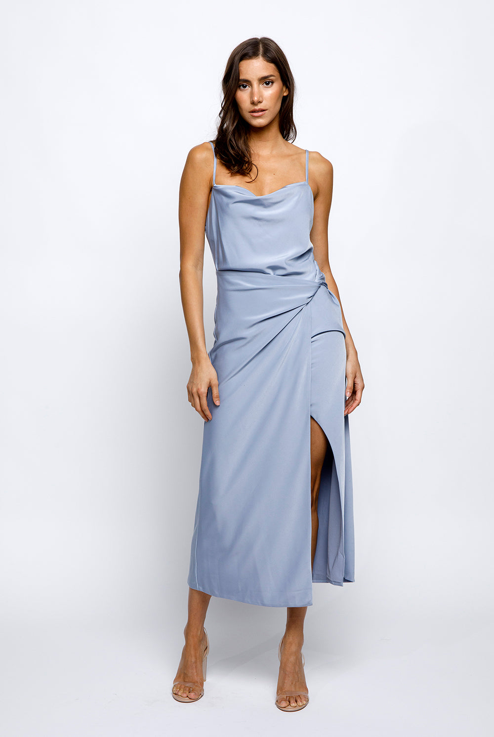 A woman wearing a blue satin slip dress with a cowlneck and adjustable spaghetti straps. The dress features a large front slit and a low open back.