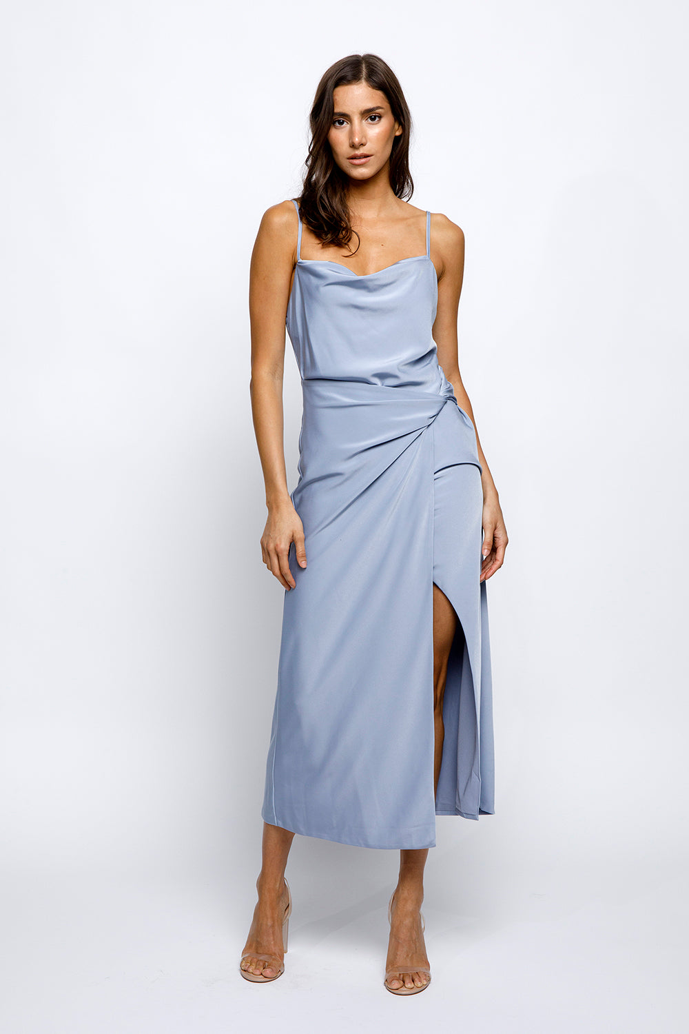 A woman wearing a blue satin slip dress with a cowlneck and adjustable spaghetti straps. The dress features a large front slit and a low open back.