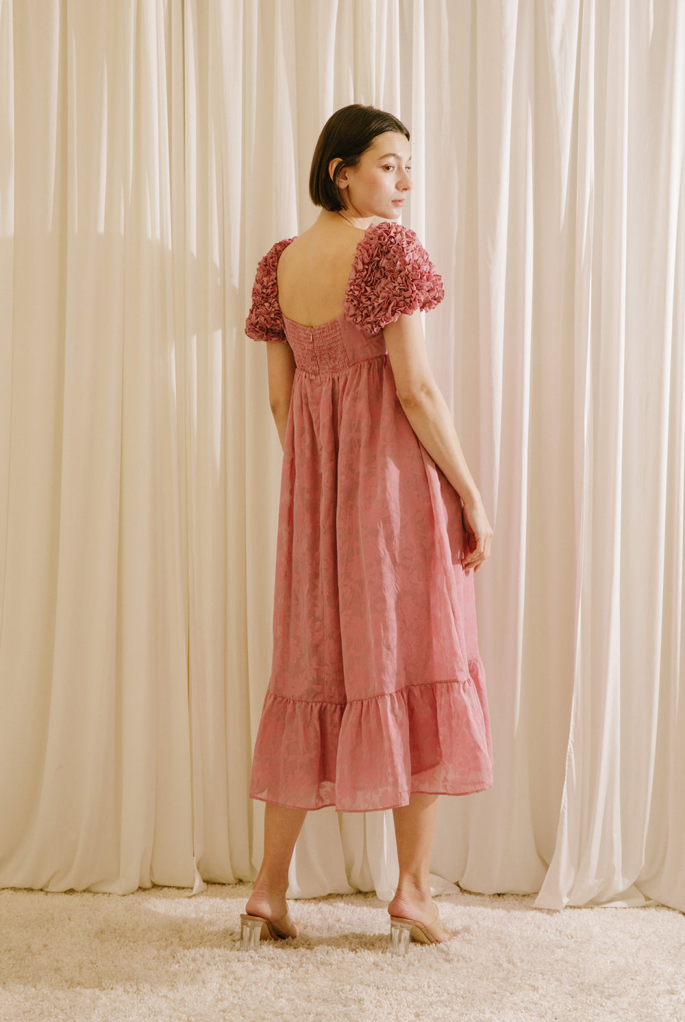 Back of a model wearing a pink dress with floral sleeves against a beige curtain backdrop