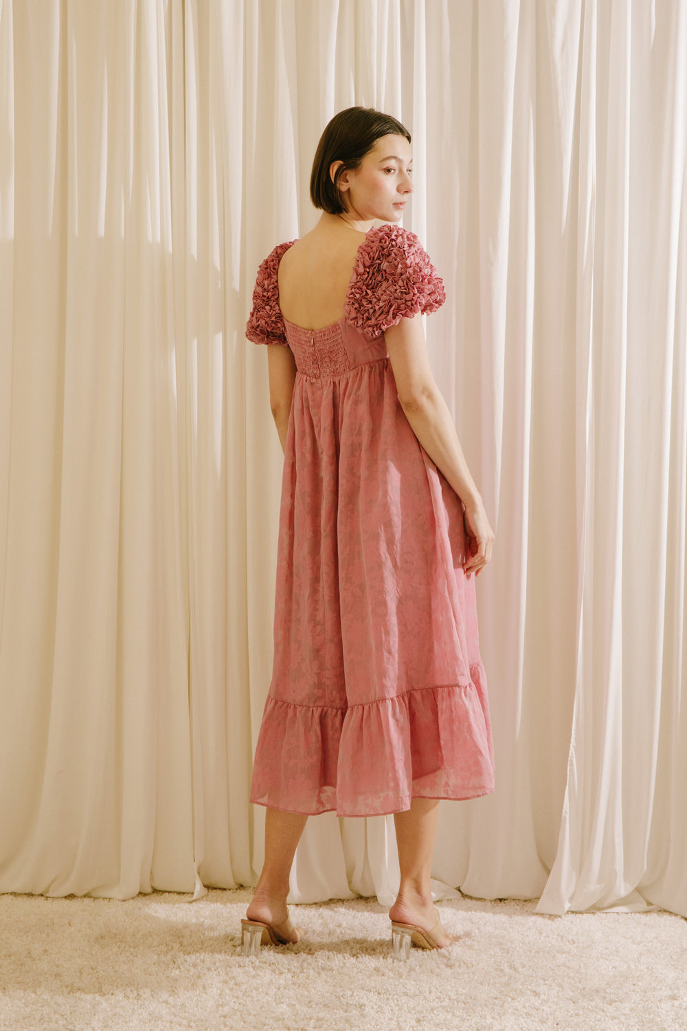 Back of a model wearing a pink dress with floral sleeves against a beige curtain backdrop