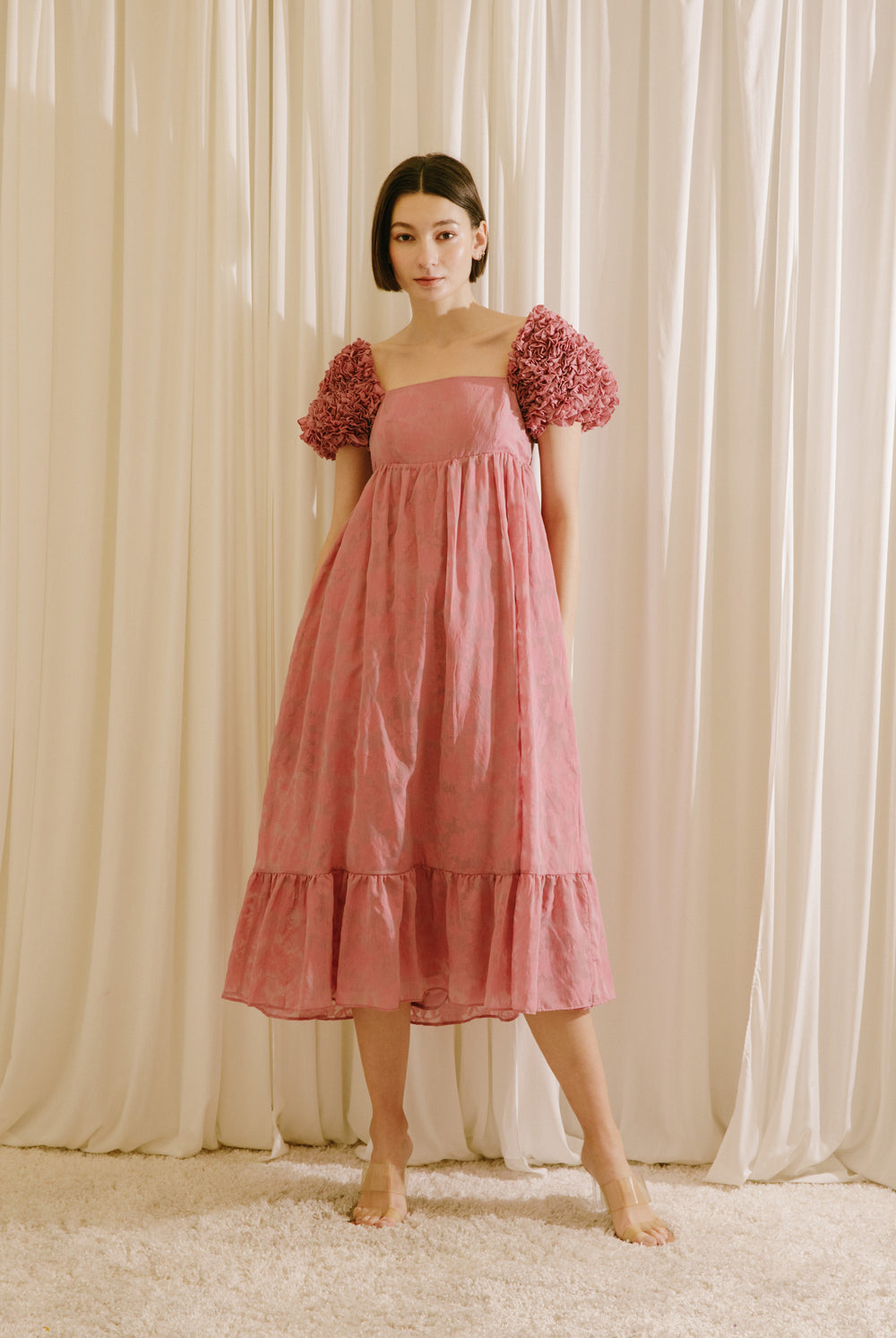 Woman wearing a pink dress with floral sleeves against a beige curtain background