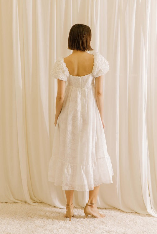 Back view of a woman wearing a white midi dress with statement rosette puff sleeves standing against a beige curtain
