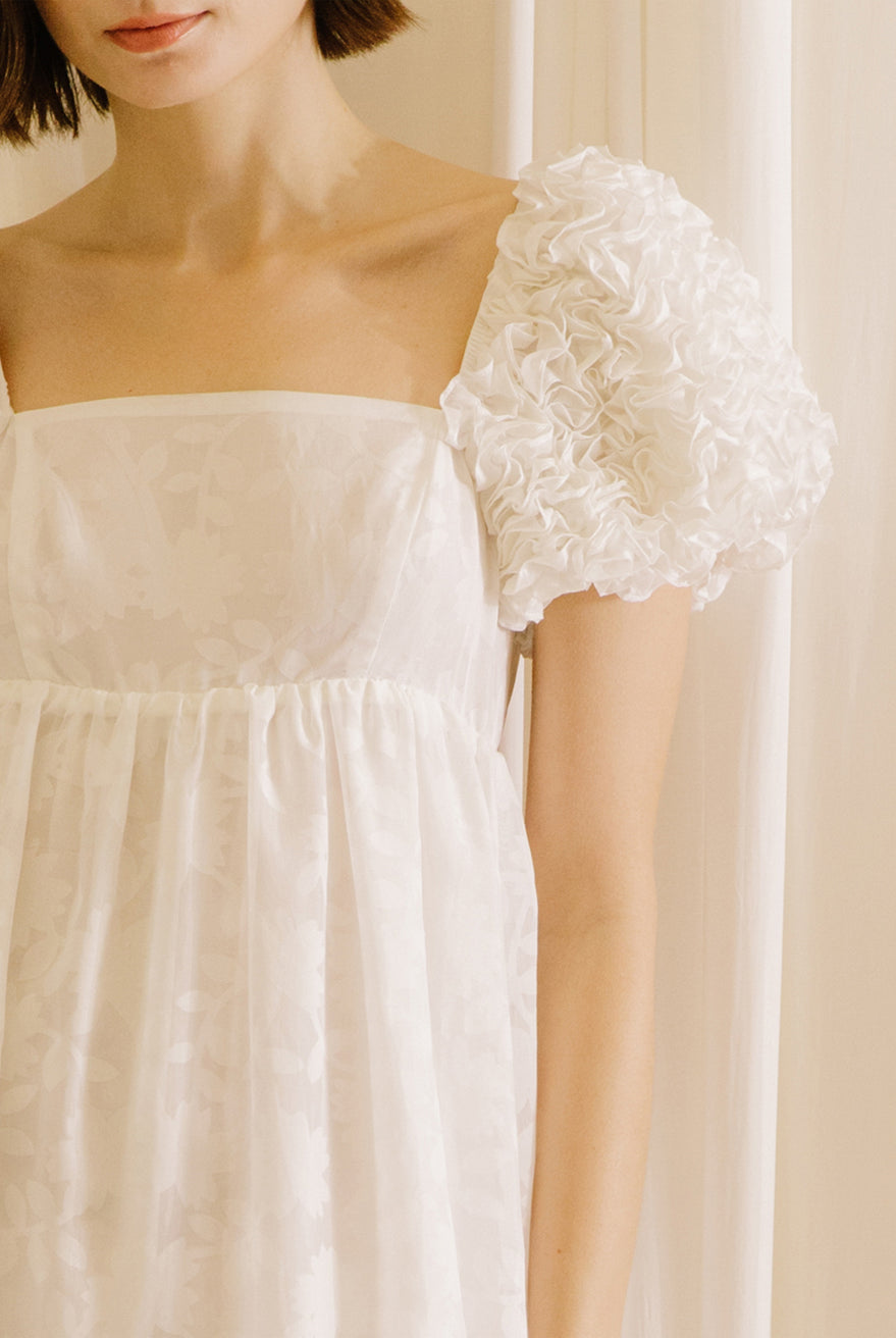 White midi dress with statement rosette puff sleeves standing against a beige curtain