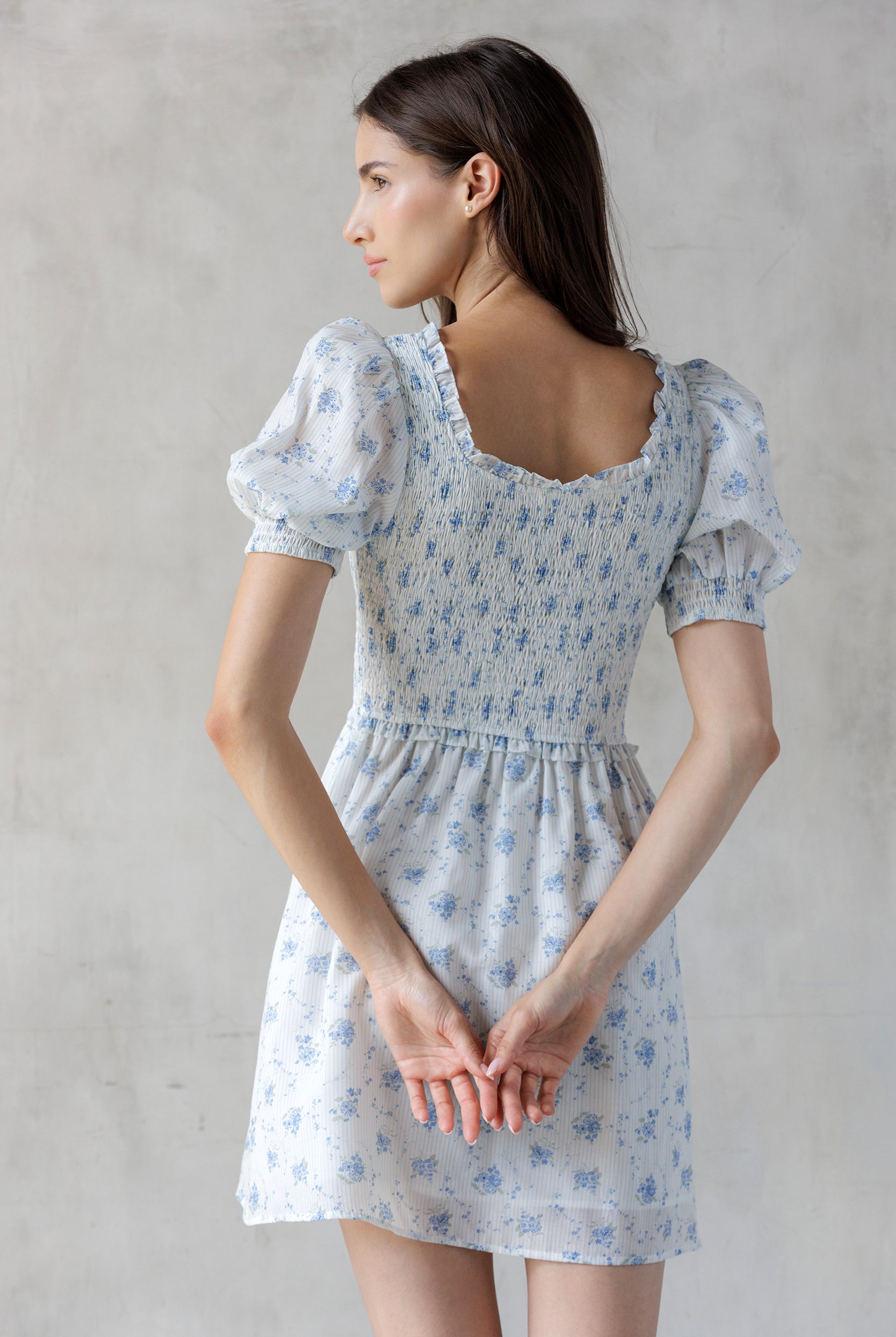 A woman viewed from behind wearing a white and striped dress with a blue floral print and a smocked bodice against a gray wall.