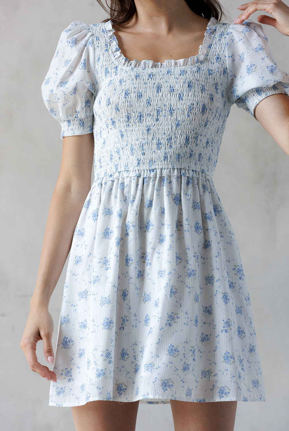 A close-up of a person wearing a white and striped dress with a blue floral print and a smocked bodice against a gray wall.
