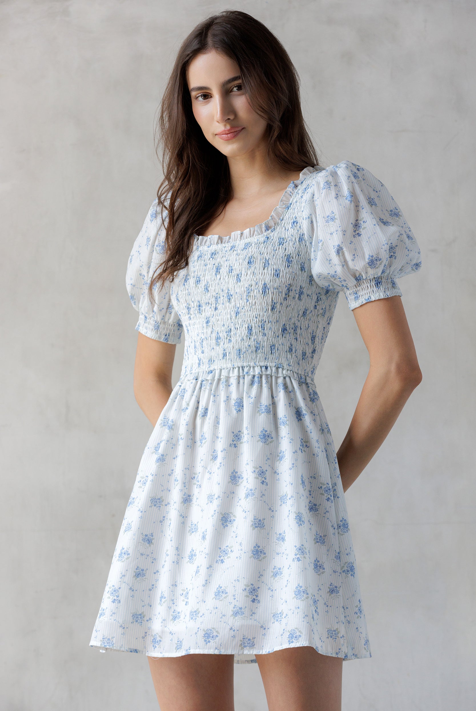 Model wearing a white and striped dress with a blue floral print and a smocked bodice against a gray wall.