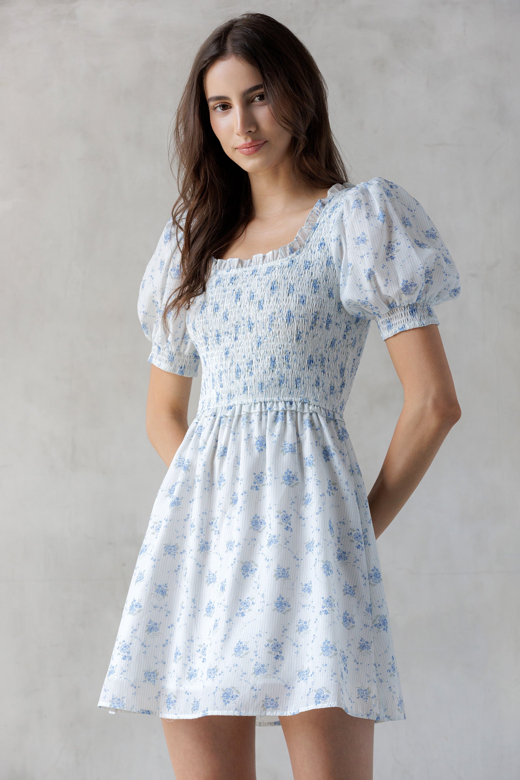 Model wearing a white and striped dress with a blue floral print and a smocked bodice against a gray wall.