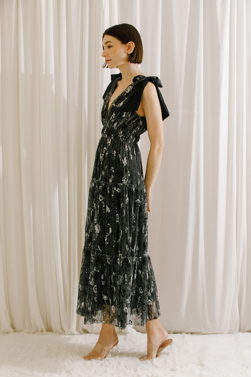 Side view of a model wearing a black and silver metallic floral dress against a white curtain background