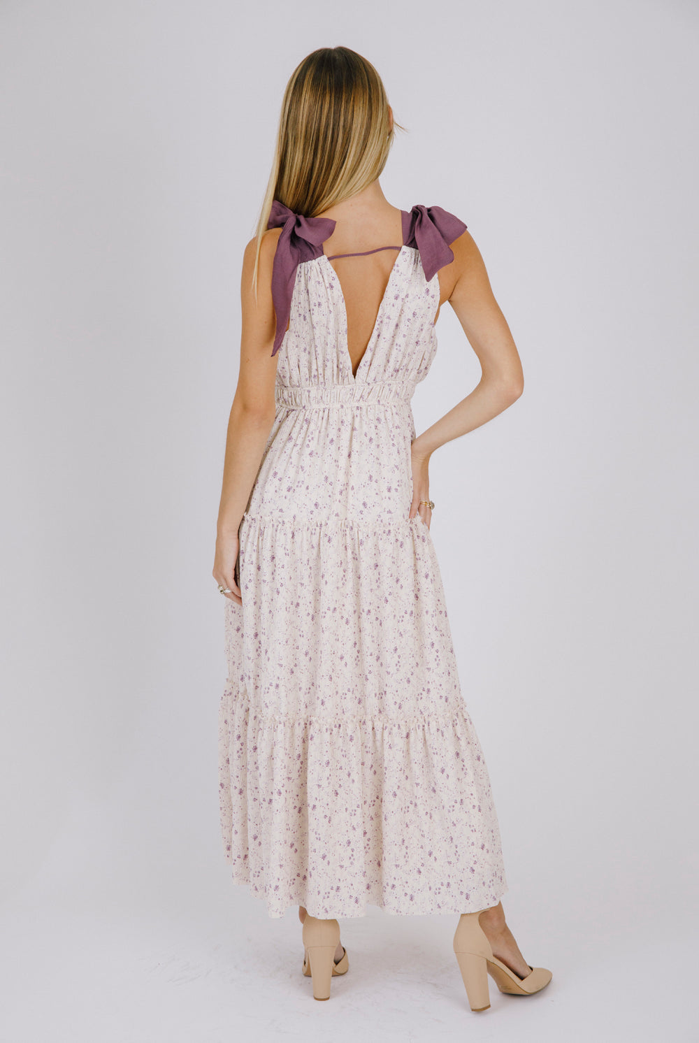 Back view of a woman wearing a purple floral maxi dress with bow straps details on a white background