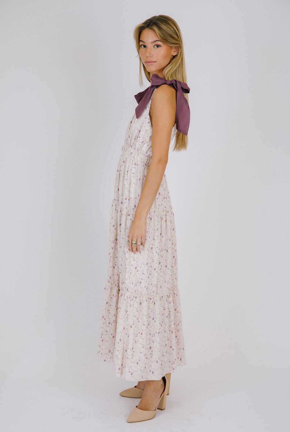 Side view of a woman wearing a purple floral maxi dress with bow straps details on a white background