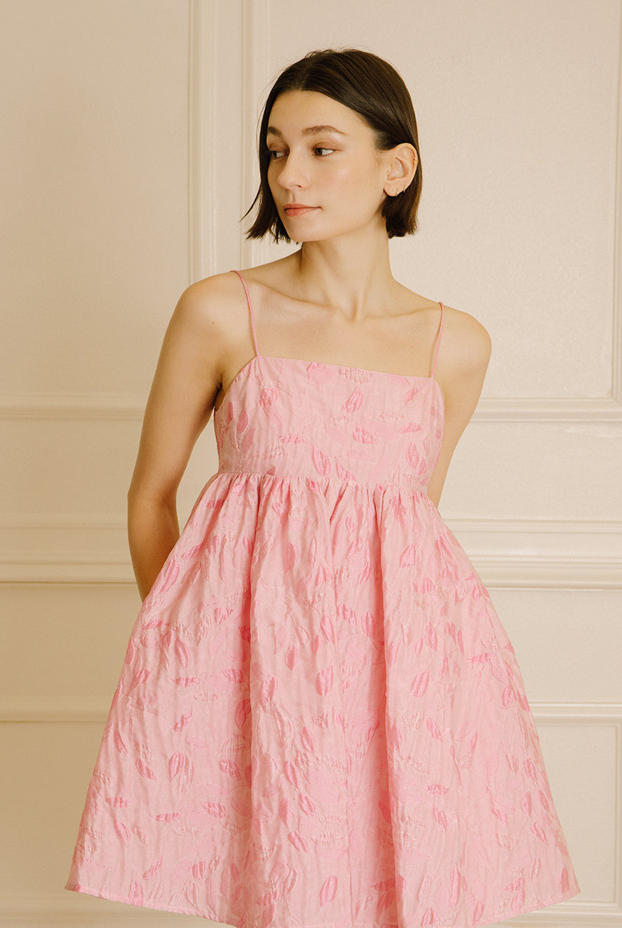 Woman wearing a pink dress with floral jacquard throughout and thin straps against a beige wall