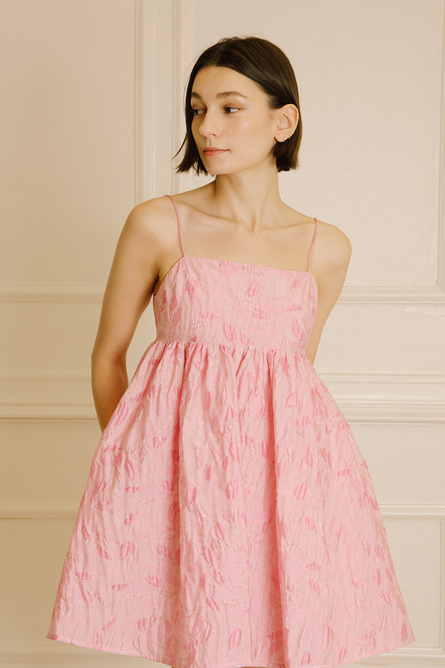 Woman wearing a pink dress with floral jacquard throughout and thin straps against a beige wall