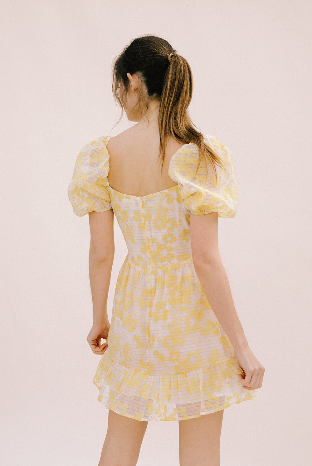 Back view of a woman wearing a yellow floral mini dress with an invisible zipper on a light pink background