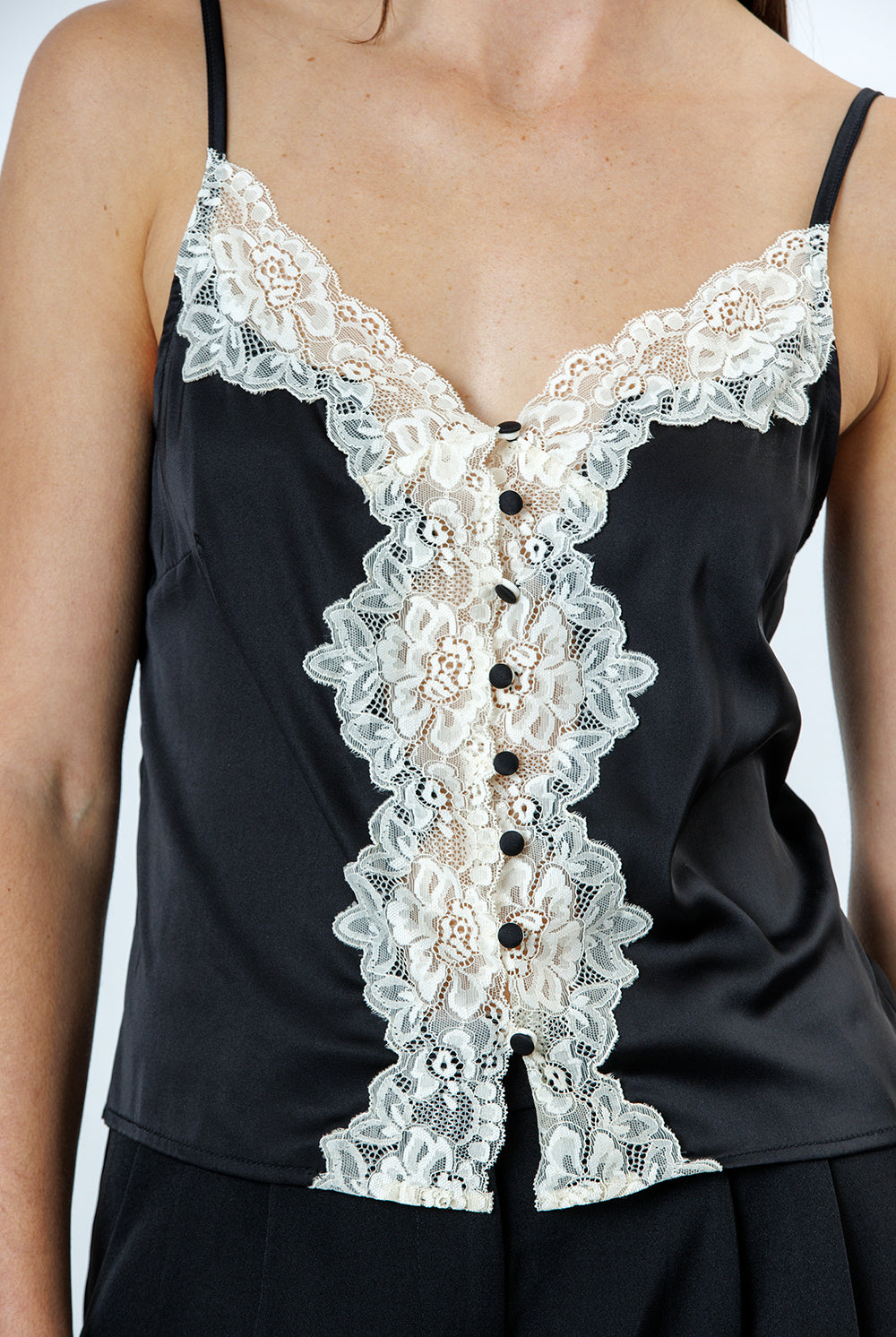 A person wearing a black top with floral lace trim, a v-neckline, adjustable spaghetti straps, and vintage-inspired button-front details.