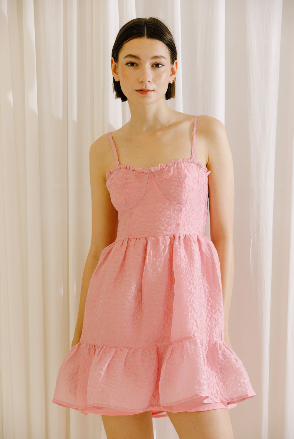 Woman wearing a pink textured dress against a white curtain background