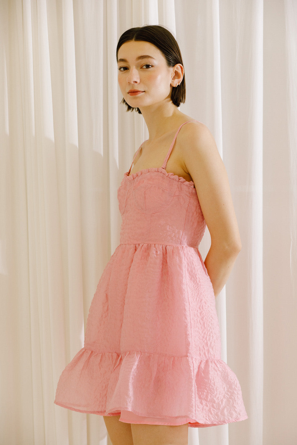 Side view of a woman wearing a pink textured dress against a white curtain background