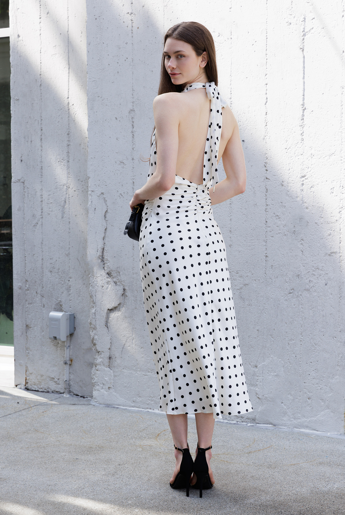 A woman stands outdoors against a textured light gray wall, wearing an ivory halter-neck midi dress with black polka dots and a large open back. She is holding a small black handbag and wearing black ankle-strap heels.