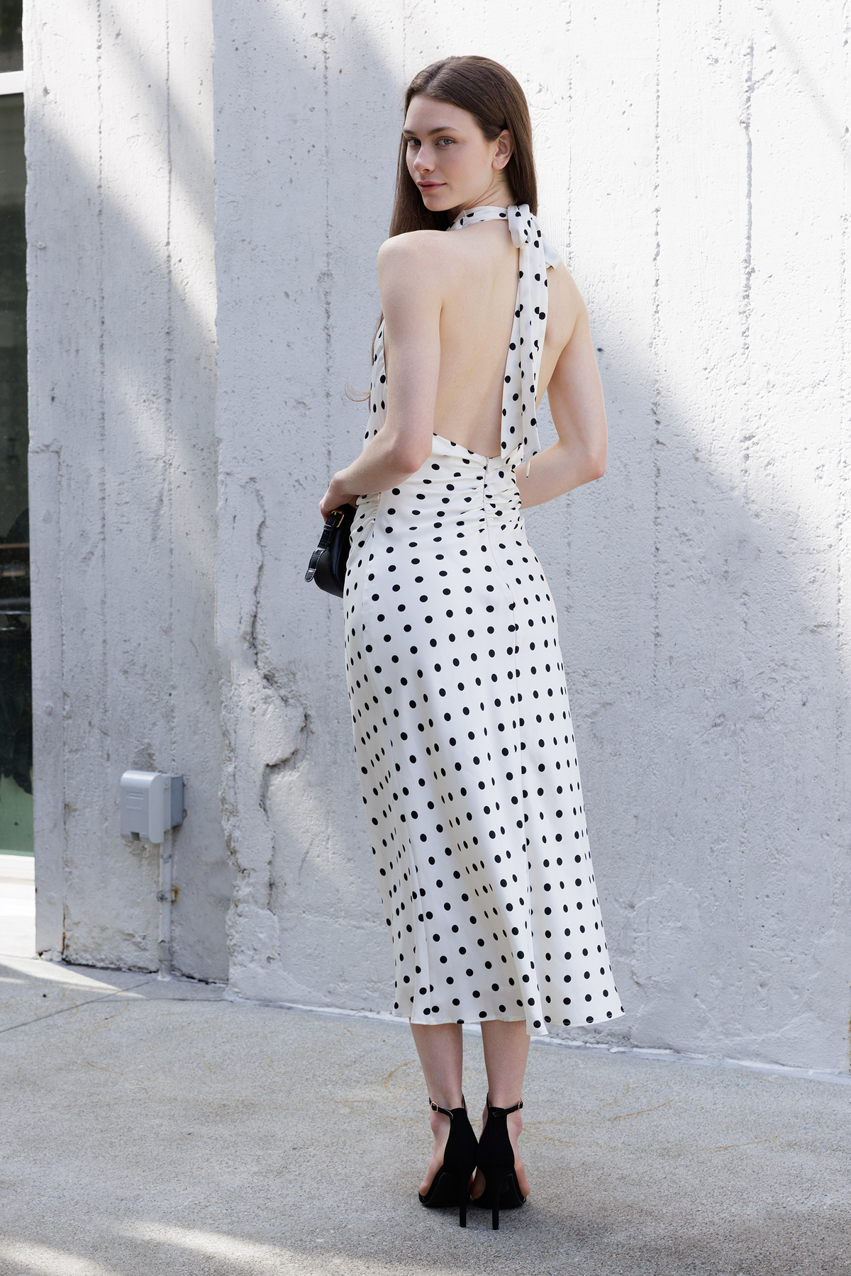 A woman stands outdoors against a textured light gray wall, wearing an ivory halter-neck midi dress with black polka dots and a large open back. She is holding a small black handbag and wearing black ankle-strap heels.