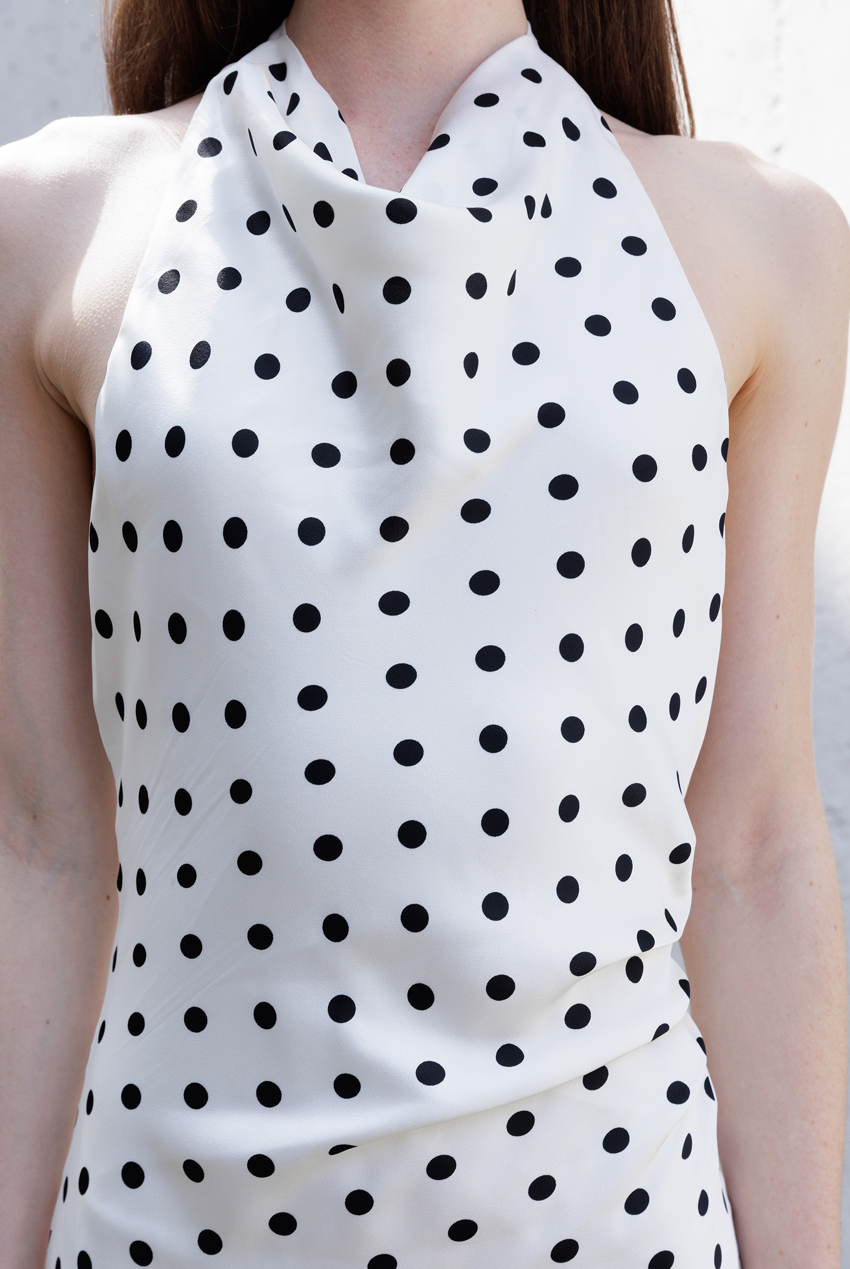 A close-up of a woman wearing a sleeveless halter-neck top or dress in ivory with an allover black polka dot pattern.