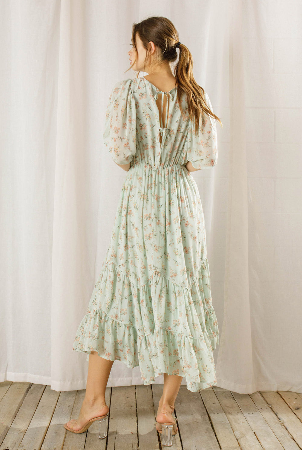 Back view of a woman wearing a sage dress with a floral pattern standing against a white curtain background