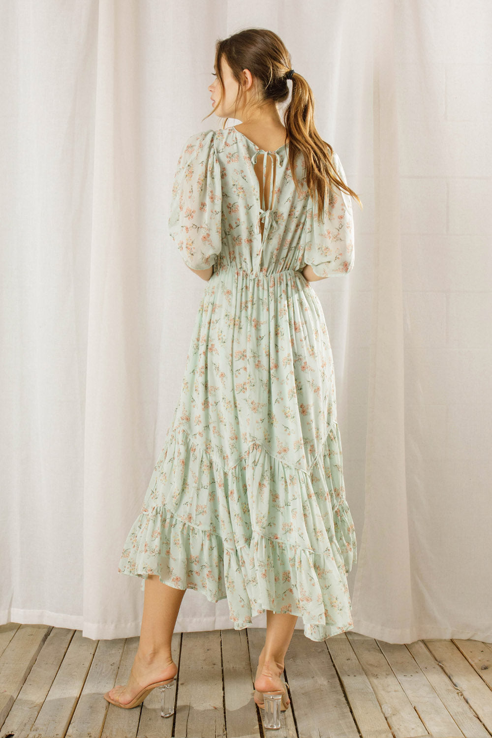 Back view of a woman wearing a sage dress with a floral pattern standing against a white curtain background