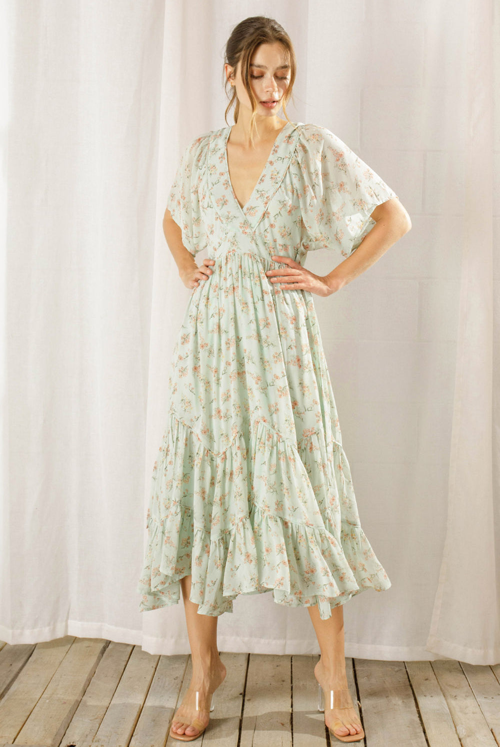Woman wearing a sage dress with a floral pattern standing against a white curtain background