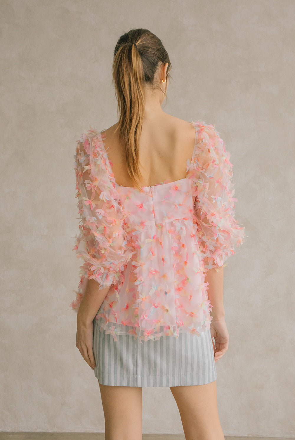 Back view of a woman wearing a pink tulle top with a blue striped skirt against a neutral wall.