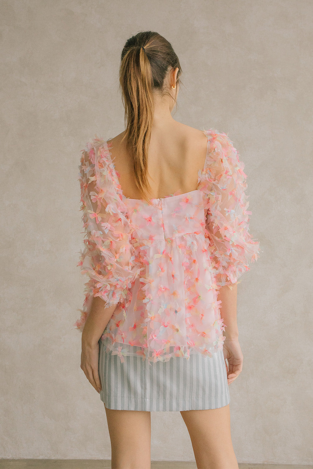 Back view of a woman wearing a pink tulle top with a blue striped skirt against a neutral wall.