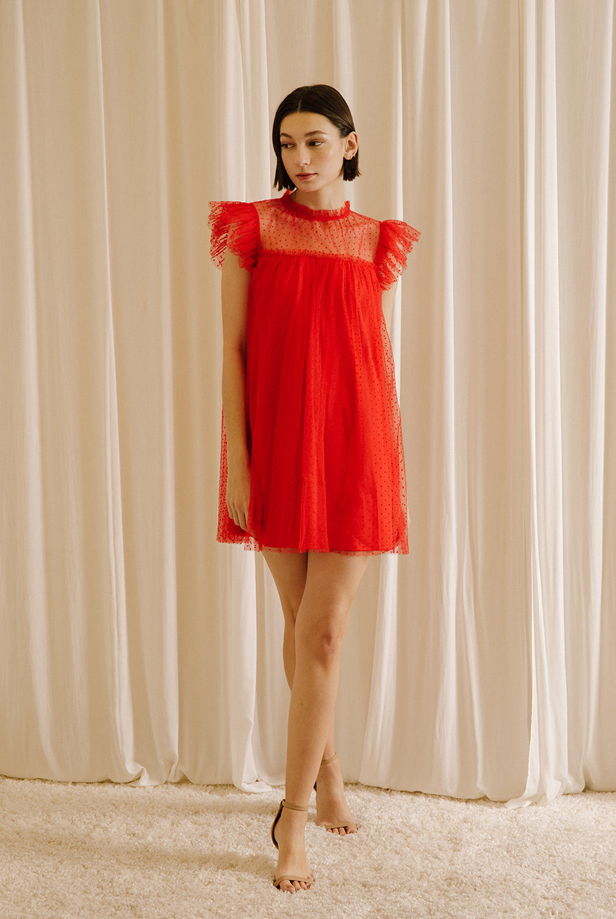Alternative view of a woman wearing a red dress with tulle fabric with mini polka dot design throughout against a beige wall.