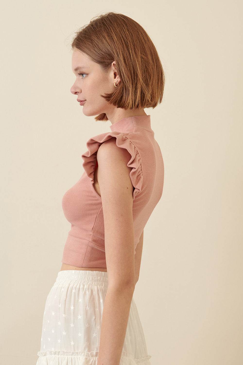 Side view of a woman wearing a pink sleeveless top with ruffled shoulders against a beige background