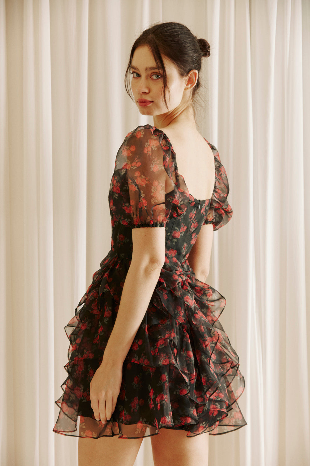 Side view of a woman wearing a black floral dress with sheer puff sleeves against a light curtain background