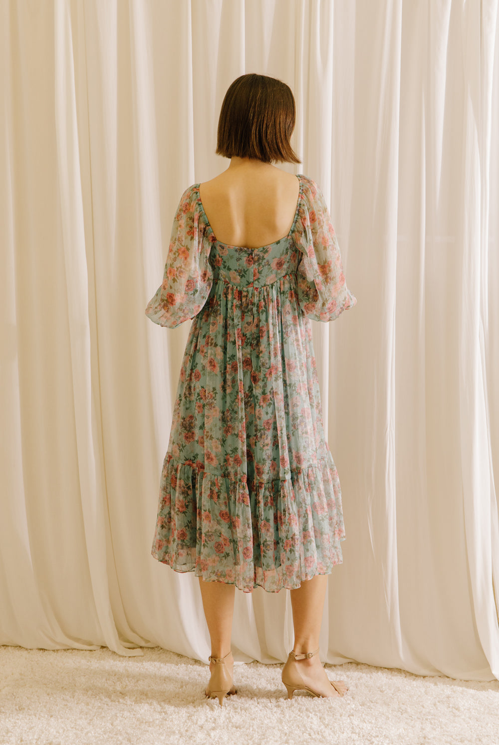Back view of a woman wearing a teal floral midi dress against a beige curtain background