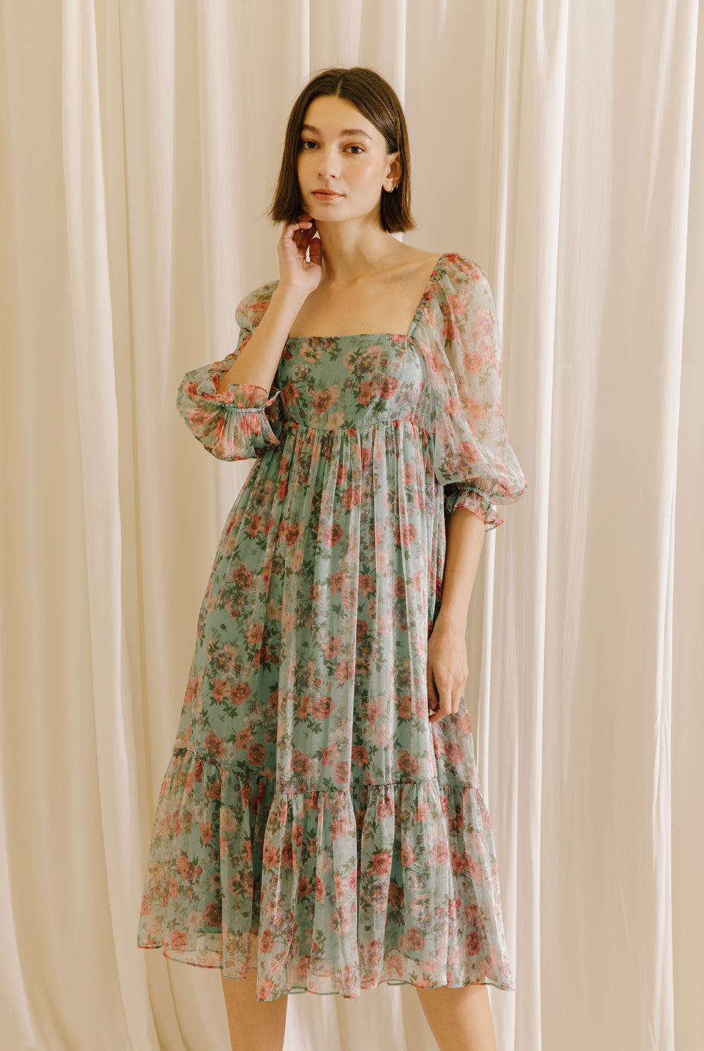 Woman wearing a teal floral midi dress against a beige curtain background