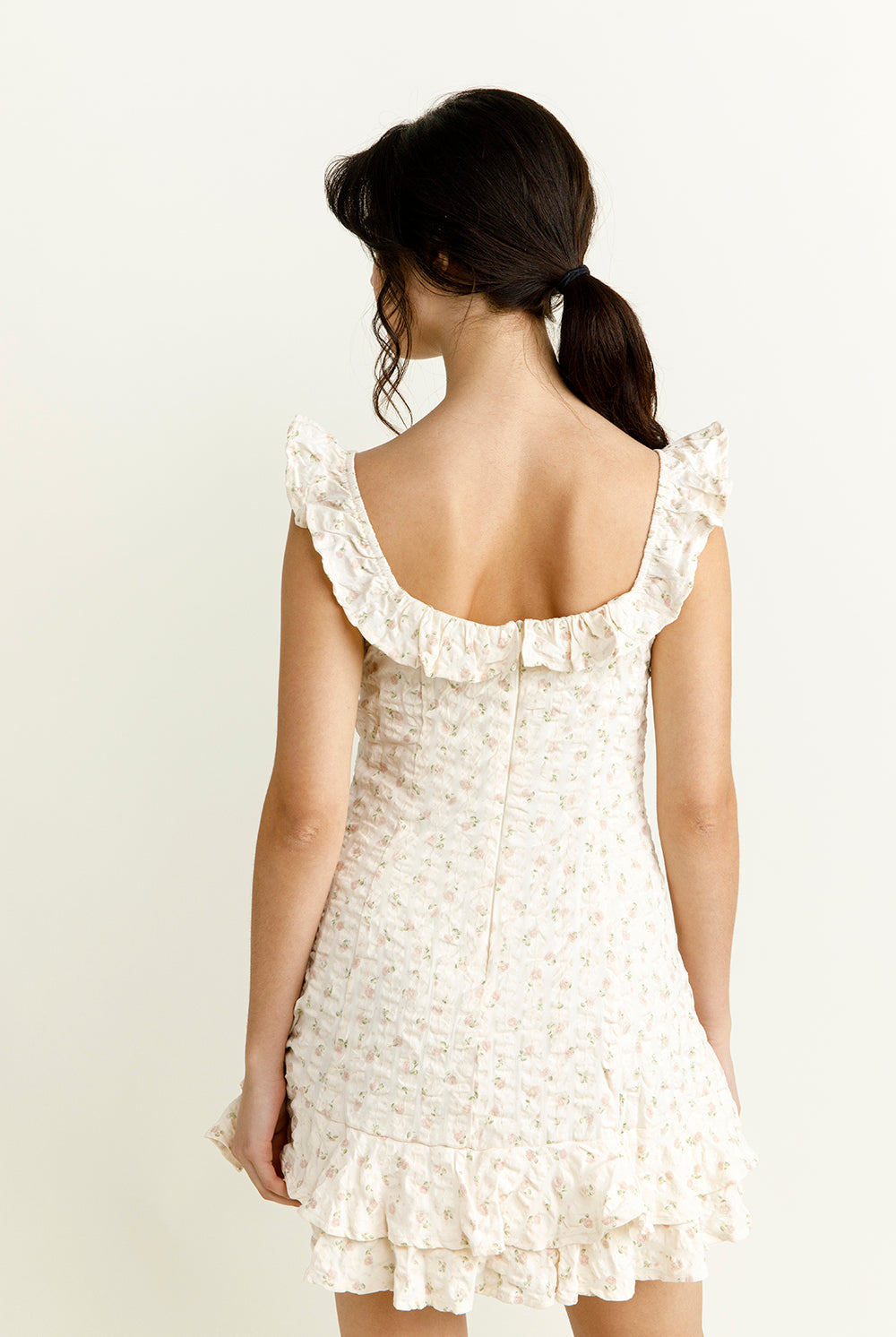 Back view of a woman wearing an ivory floral mini dress with ruffled straps against a plain background