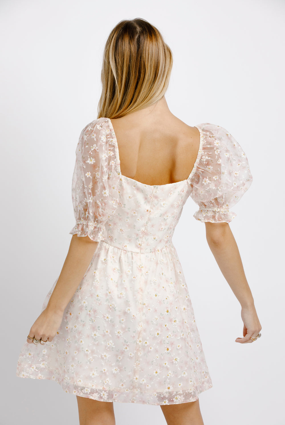Back view of woman wearing a cream dress with floral pattern and sheer puffy sleeves on a plain background