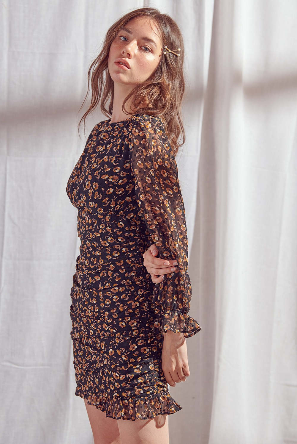 A woman modeling a long-sleeve black mini dress with a orange and yellow floral print pattern, posed against a draped white fabric backdrop.