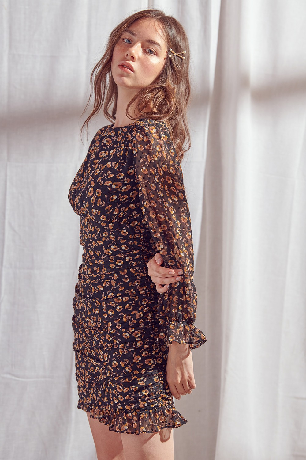 A woman modeling a long-sleeve black mini dress with a orange and yellow floral print pattern, posed against a draped white fabric backdrop.