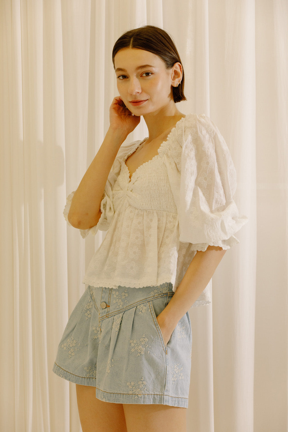 A woman modeling a white embroidered puff-sleeve blouse and light blue denim shorts against a soft cream curtain background.