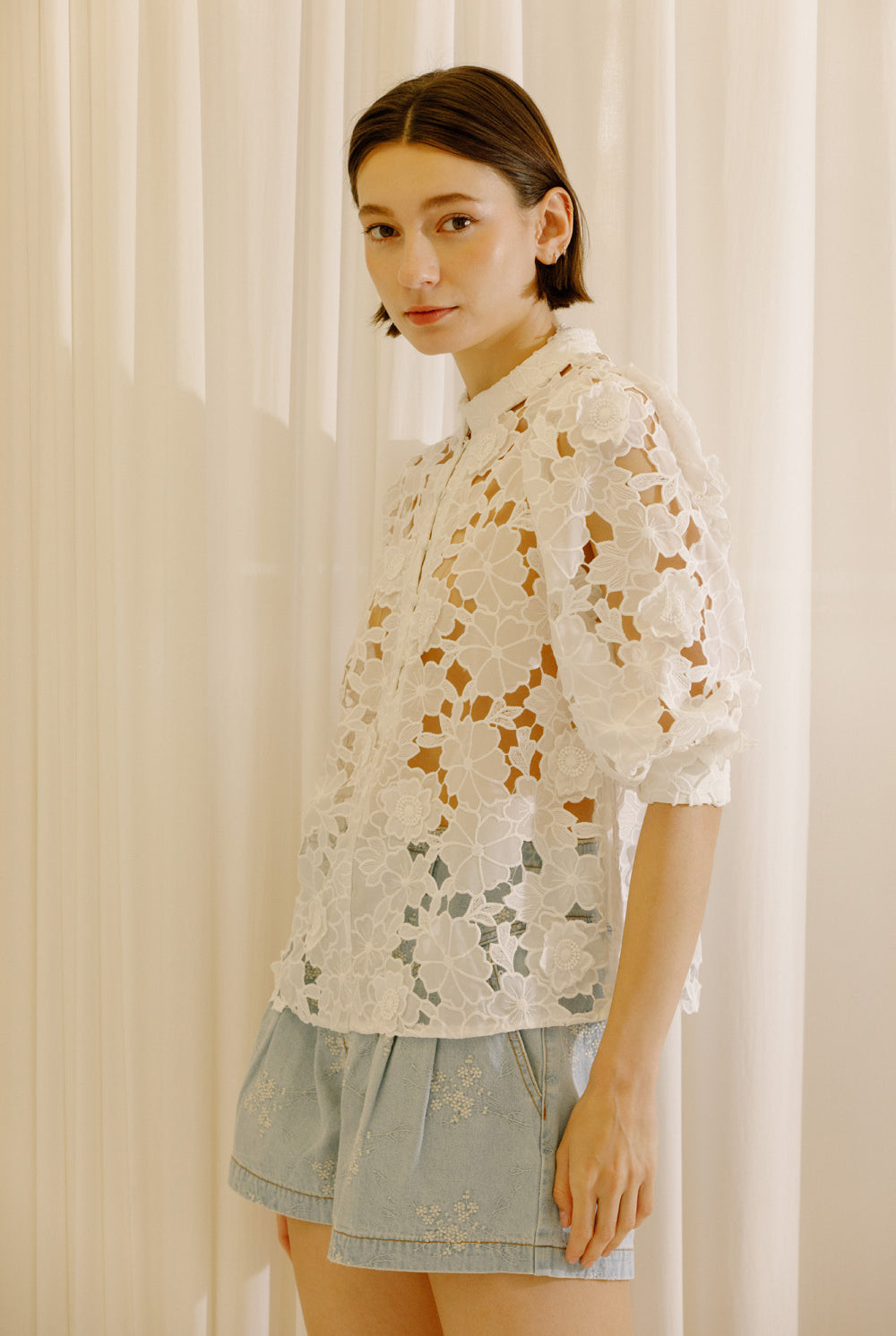 A woman wearing a white eyelet lace blouse with puff sleeves and light blue denim shorts, standing in front of a soft beige curtain backdrop.