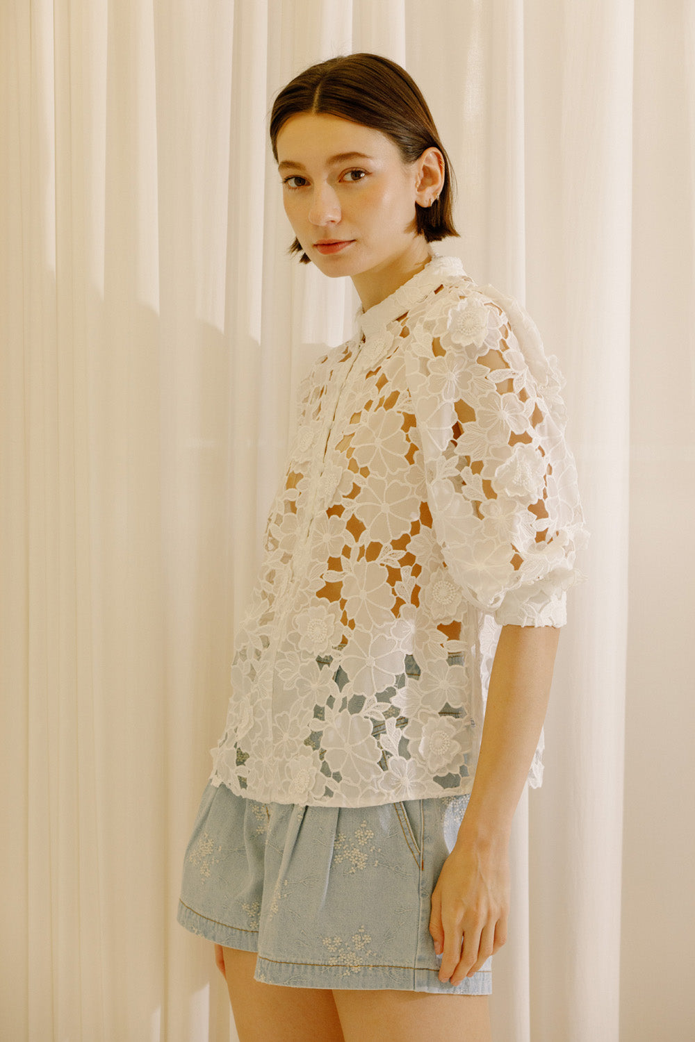 A woman wearing a white eyelet lace blouse with puff sleeves and light blue denim shorts, standing in front of a soft beige curtain backdrop.