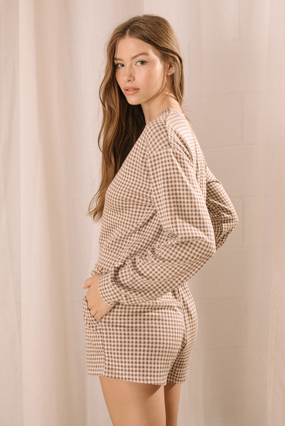 Side view of a woman modeling a lounge set that features a long-sleeve top and high-waisted drawstring shorts with pockets in a brown and ivory gingham print.