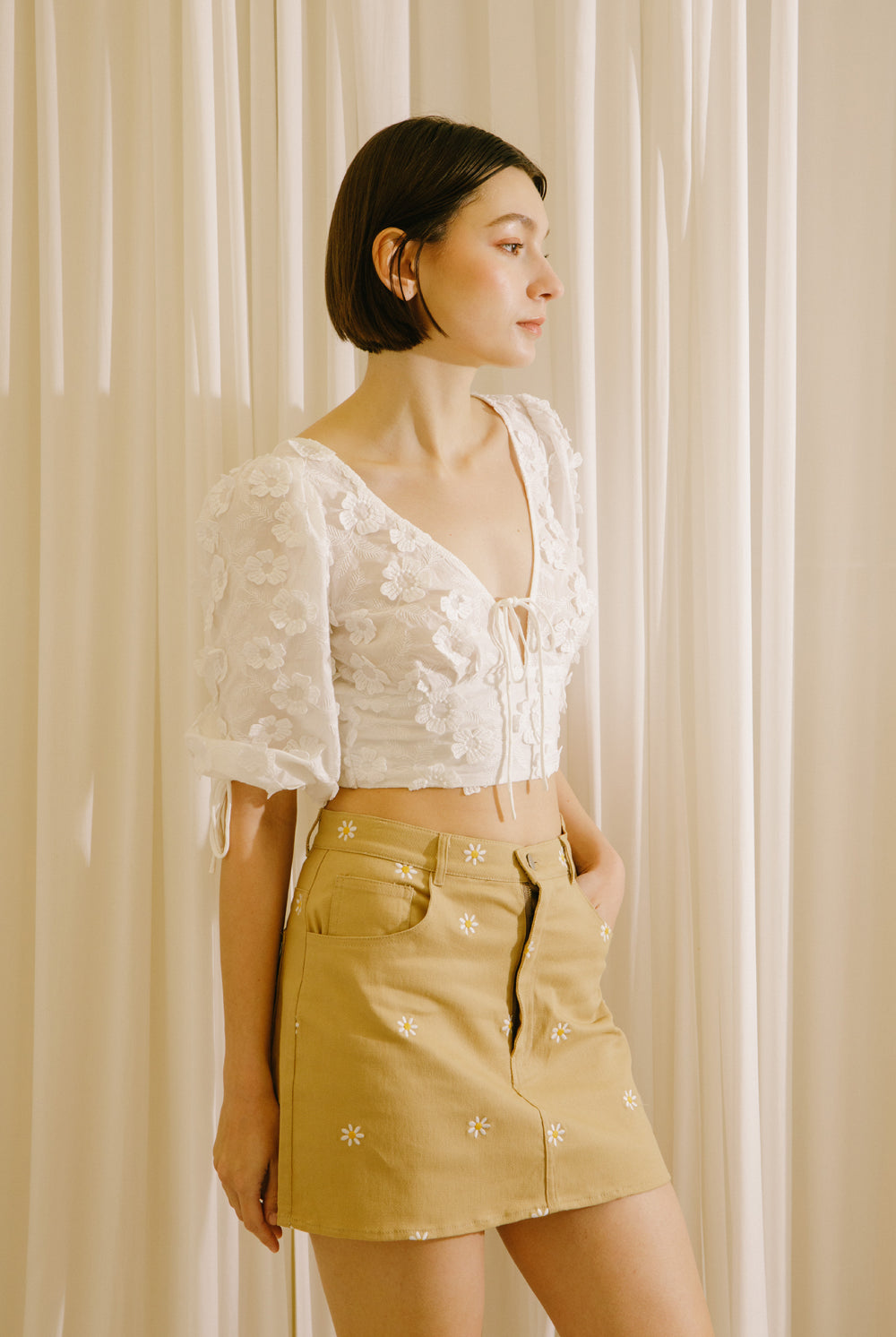 A woman wearing a white crop top with dimensional flower embellishment short-sleeve and a mustard yellow mini skirt with white embroidered details, standing in front of a cream-colored curtain backdrop.